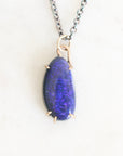 Dark colored irregularly shaped opal set in gold prongs against a plain background.