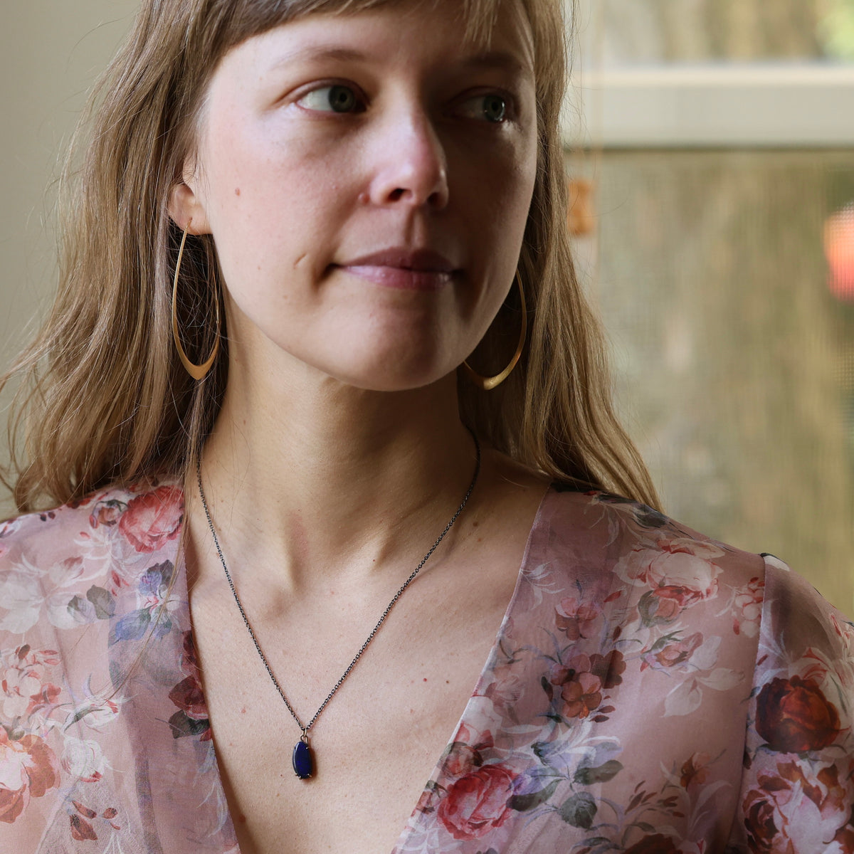 Woman wearing irregular shaped opal necklace, floral top, and large gold hoops. Indoor background. 