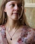 Woman wearing irregular shaped opal necklace, floral top, and large gold hoops. Indoor background. 