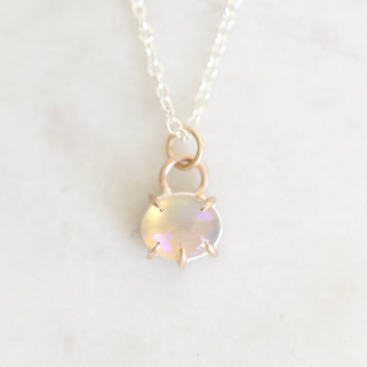 Light colored opal set in gold with a silver chain up close against a white marble background. 
