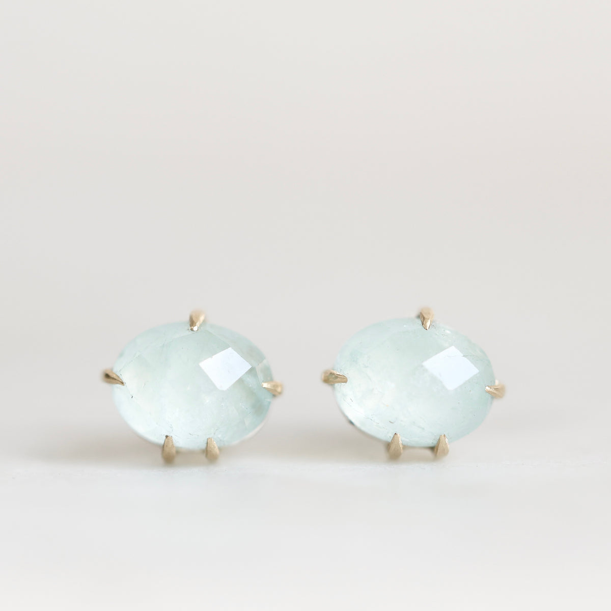 A pair of oval stud earrings featuring light blue-green cabochon gemstones in a gold prong setting, displayed against a clean, light off-white background.