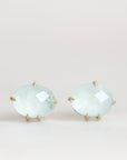 A pair of oval stud earrings featuring light blue-green cabochon gemstones in a gold prong setting, displayed against a clean, light off-white background.