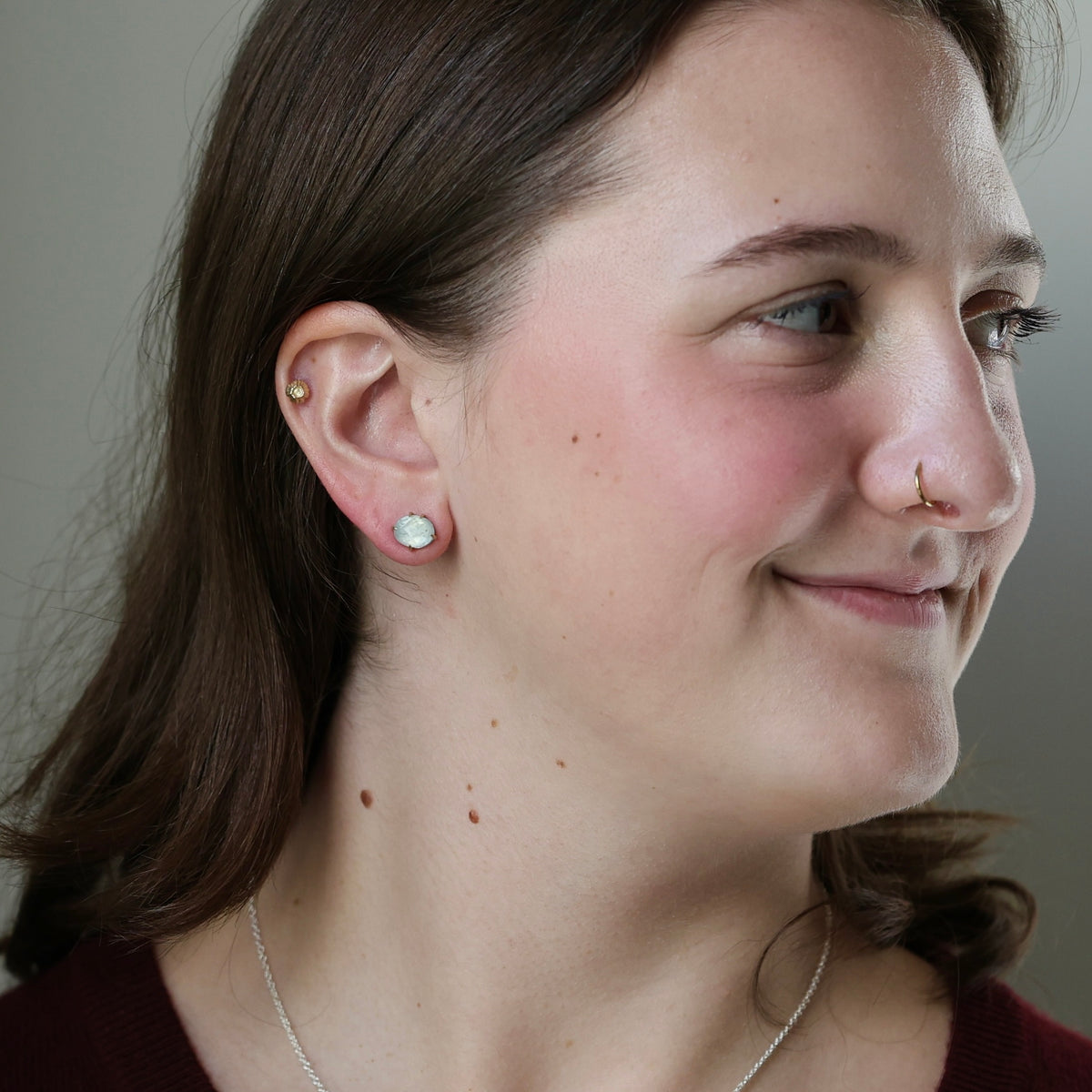 A close-up of a woman's ear wearing a small round silver stud earring with a clear gemstone, along with other ear piercings. The background is a soft, neutral gray.