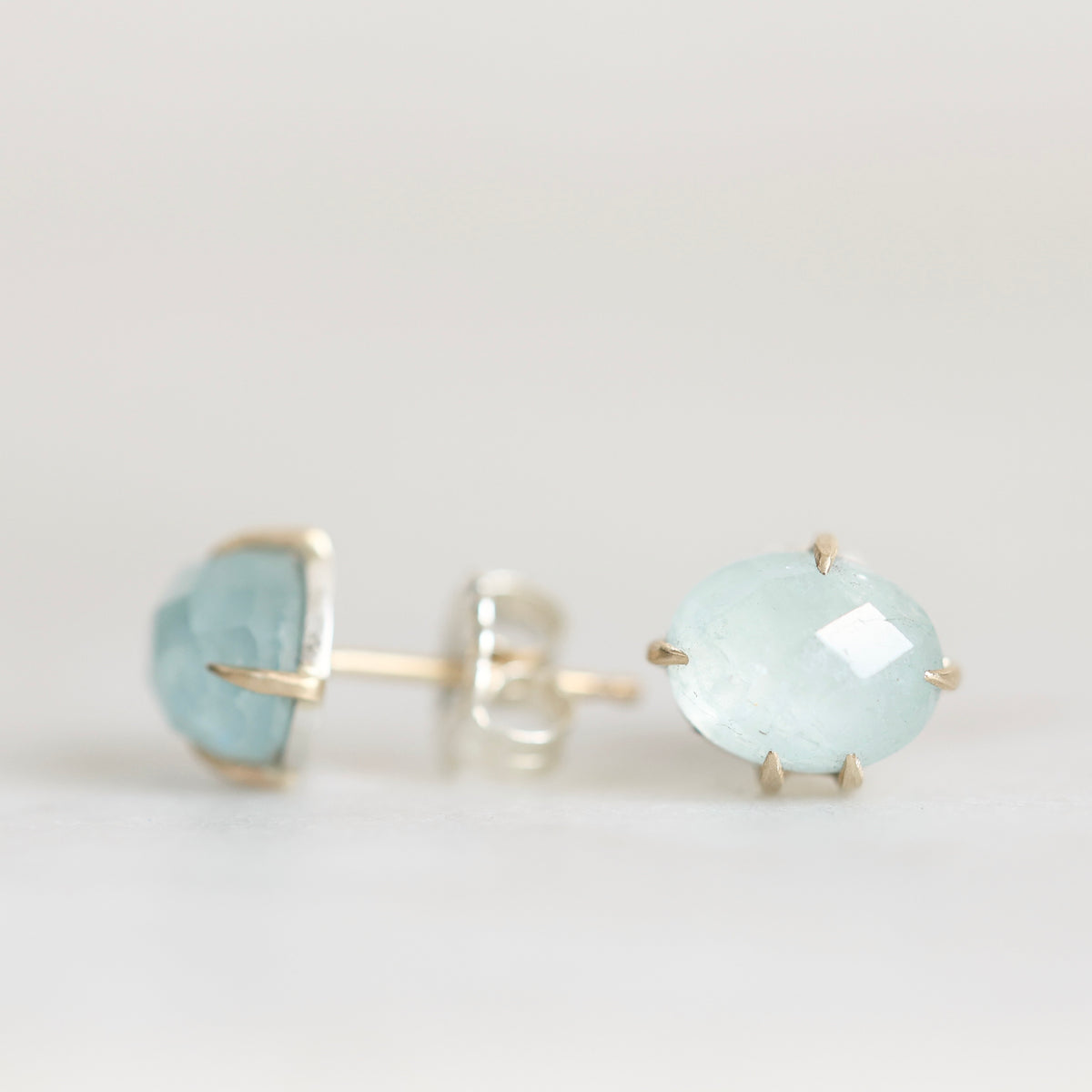 A pair of oval-cut light blue gemstone stud earrings in gold settings, with one earring shown from the front and the other from the side/back, on a plain light background.