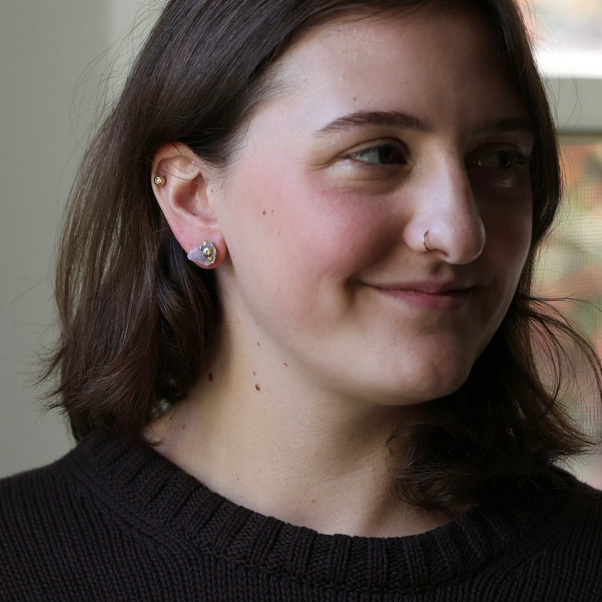 A close-up portrait of a woman with brown hair wearing a small silver hoop earring with a clear gemstone, shown from the chest up against a softly blurred indoor background.