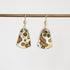 A pair of gold-tone dangle earrings with shield-shaped resin pendants featuring a white, black, and orange spotted pattern, hanging from a gold bar on a light gray background.