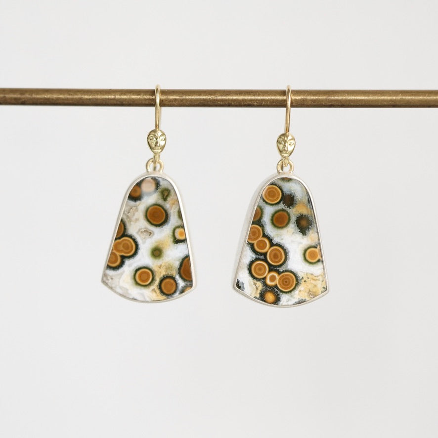 A pair of gold-tone dangle earrings with shield-shaped resin pendants featuring a white, black, and orange spotted pattern, hanging from a gold bar on a light gray background.