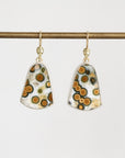 A pair of gold-tone dangle earrings with shield-shaped resin pendants featuring a white, black, and orange spotted pattern, hanging from a gold bar on a light gray background.