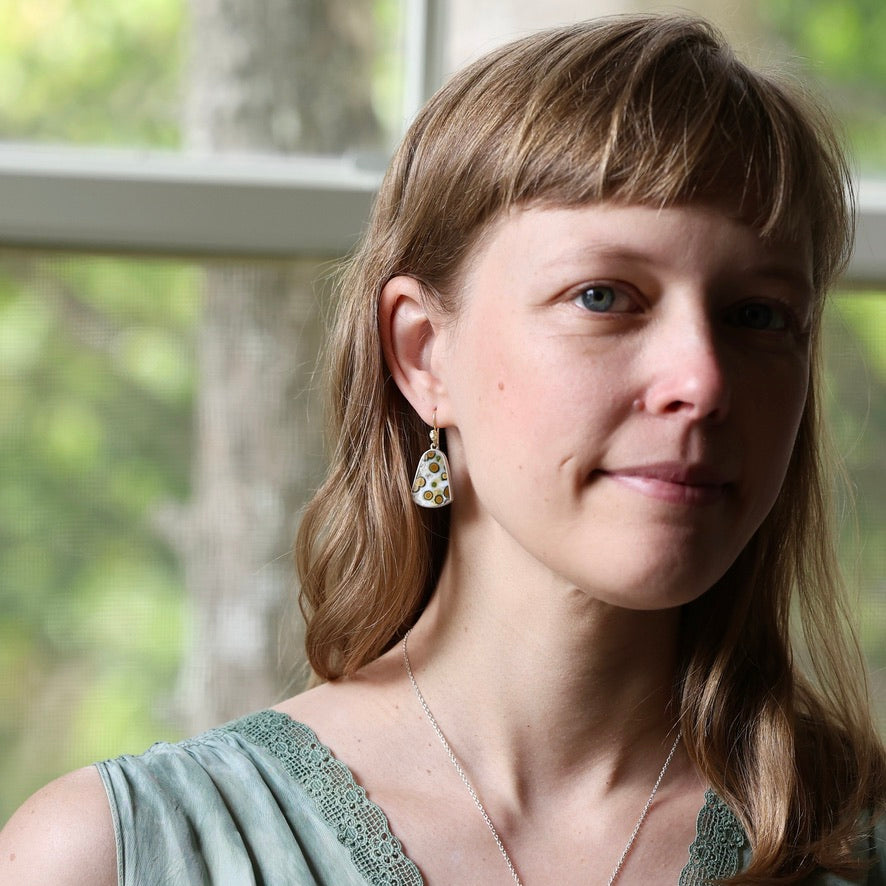 A woman with light brown hair and bangs wearing a gold teardrop earring with a white and green patterned stone, photographed in soft natural light near a window.