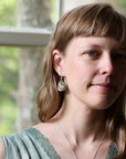 A woman with light brown hair and bangs wearing a gold teardrop earring with a white and green patterned stone, photographed in soft natural light near a window.