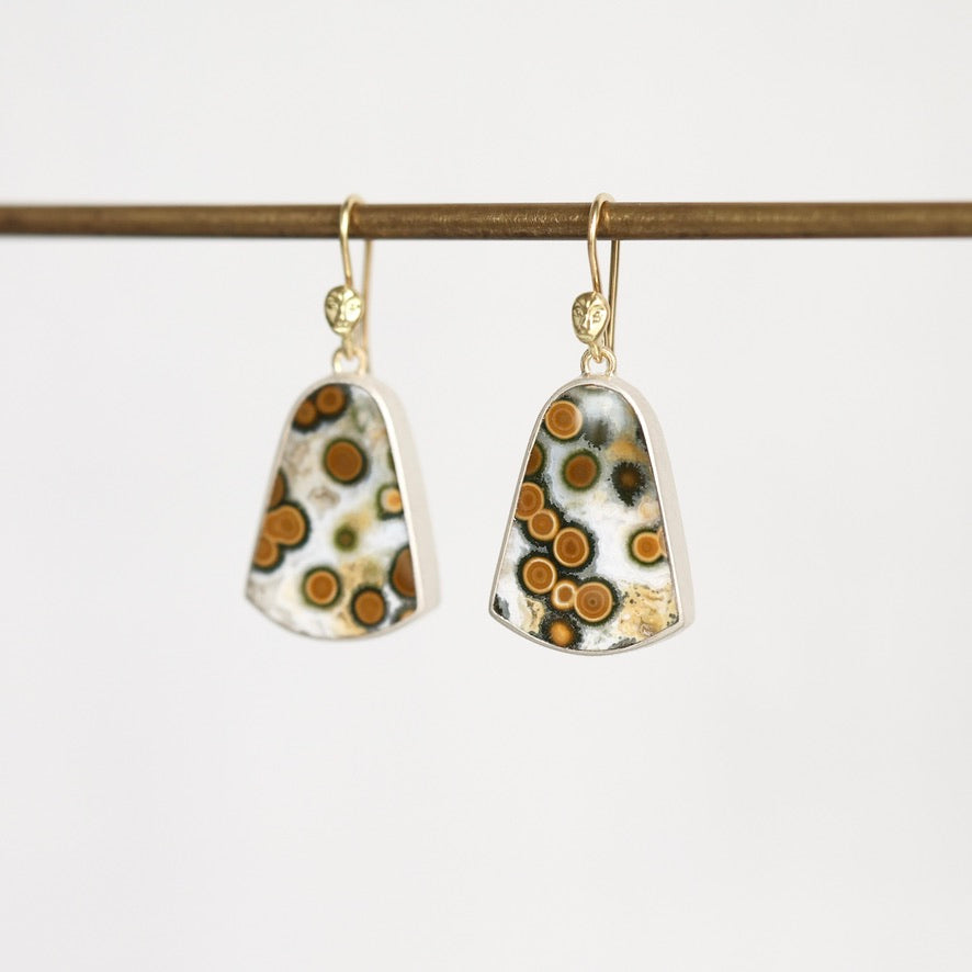 A pair of gold-tone dangle earrings with irregular shield-shaped white glass pendants featuring a pattern of brown, orange, and black spots, hanging from a dark brown rod against a white background.