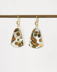 A pair of gold-tone dangle earrings with irregular shield-shaped white glass pendants featuring a pattern of brown, orange, and black spots, hanging from a dark brown rod against a white background.