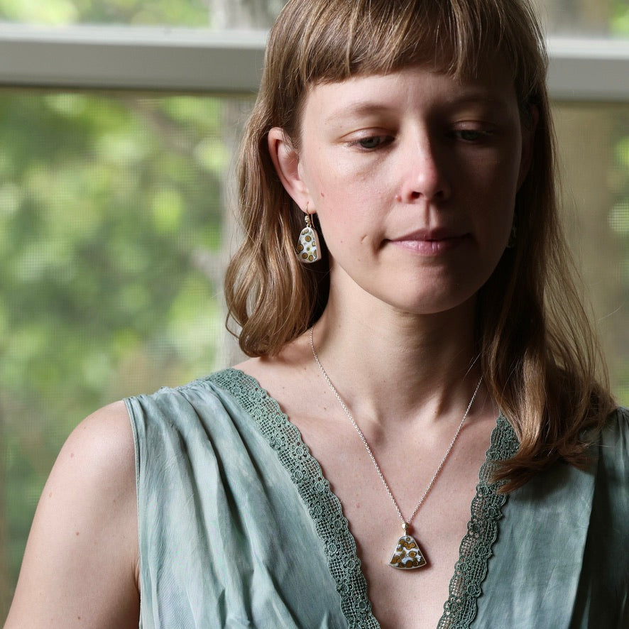 A woman wearing a silver-toned triangle pendant necklace and matching drop earrings with green and gold inlay, styled with a sage green lace-trimmed top.