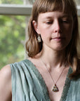 A woman wearing a silver-toned triangle pendant necklace and matching drop earrings with green and gold inlay, styled with a sage green lace-trimmed top.