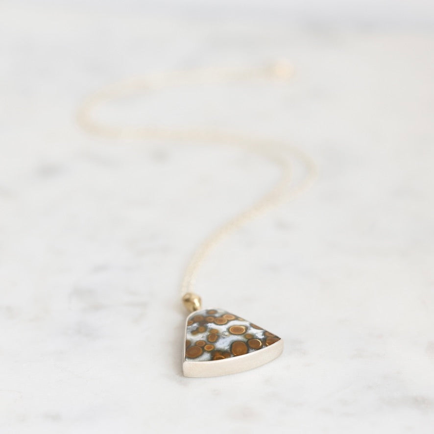 A silver-toned triangle pendant necklace with a textured, speckled brown and white pattern, suspended on a delicate gold-toned chain over a light marble background.