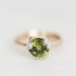 A close-up shot of a ring featuring a round green gemstone in a gold bezel setting, accented with three small clear stones on the sides, set against a plain light gray background.