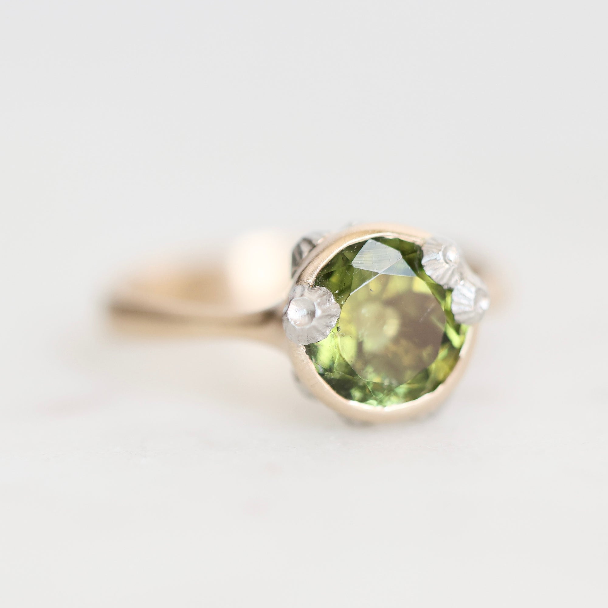 A close-up shot of a ring featuring a round green peridot gemstone in a gold setting, set against a plain light gray background.