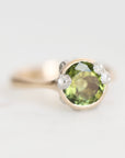 A close-up shot of a ring featuring a round green peridot gemstone in a gold setting, set against a plain light gray background.