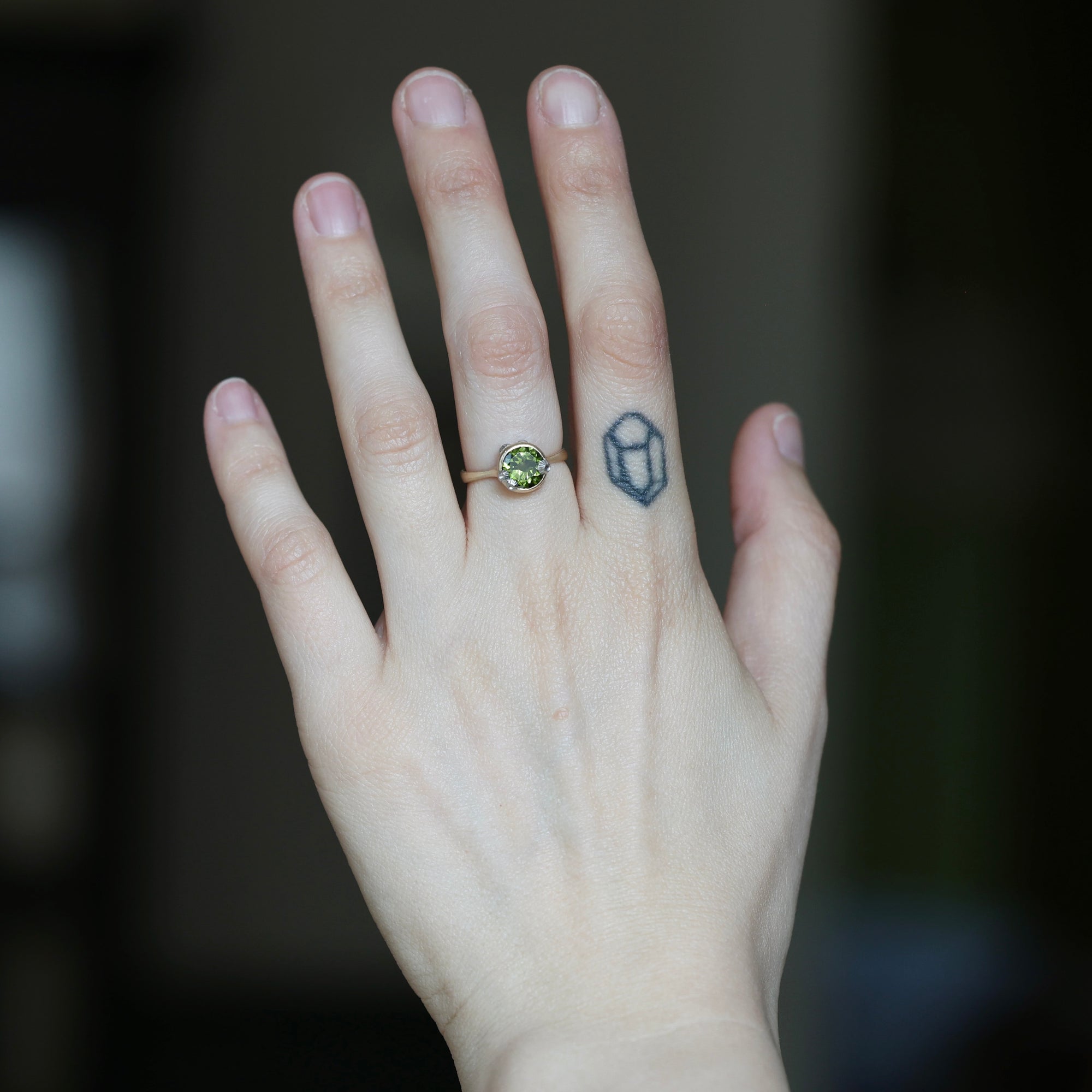 A close-up of a person's hand wearing a gold ring with a round green gemstone, set against a dark, blurred background.