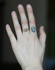 A close-up of a person's hand wearing a gold ring with a round green gemstone, set against a dark, blurred background.