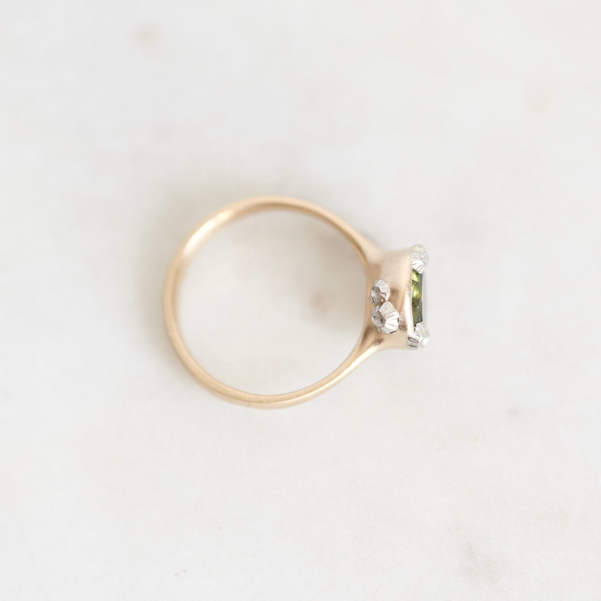 A gold ring with a small green rectangular gemstone and clear accent stones, photographed on a light marble surface.