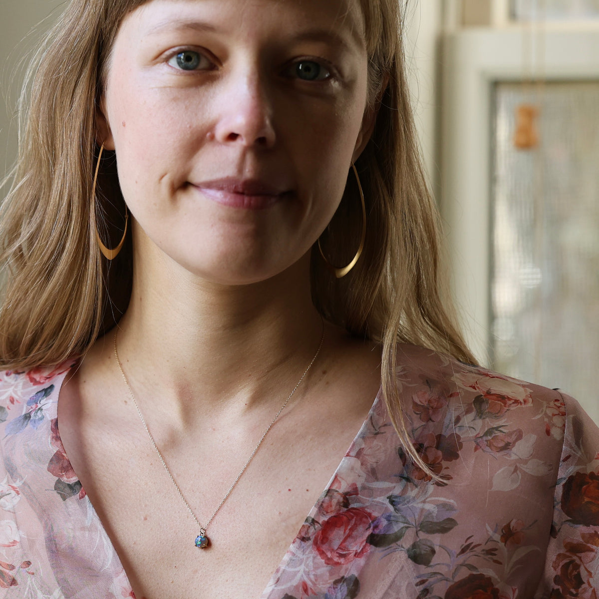Woman wearing opal necklace with gold chain. She is wearing a floral top and large gold hoops. Indoor background. 