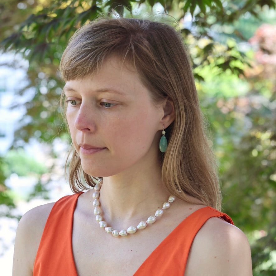 Woman wearing a pearl necklace and orange top with greenery in the background