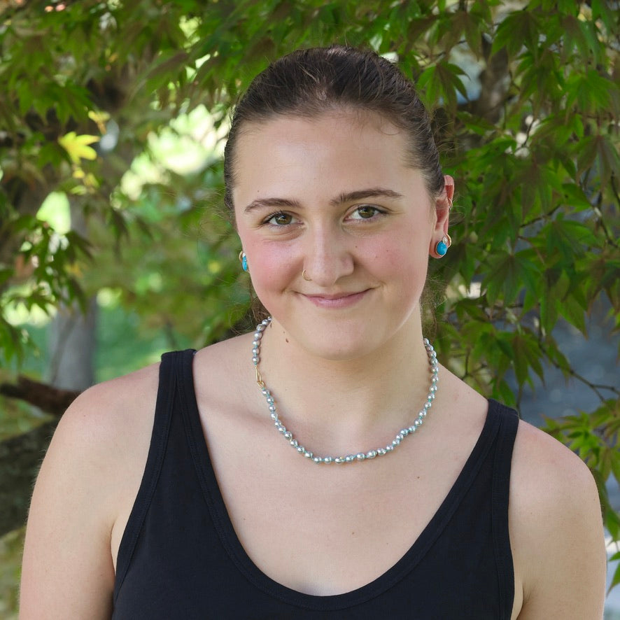 Woman wearing a black tank top and pearl necklace with greenery in the background