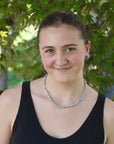 Woman wearing a black tank top and pearl necklace with greenery in the background