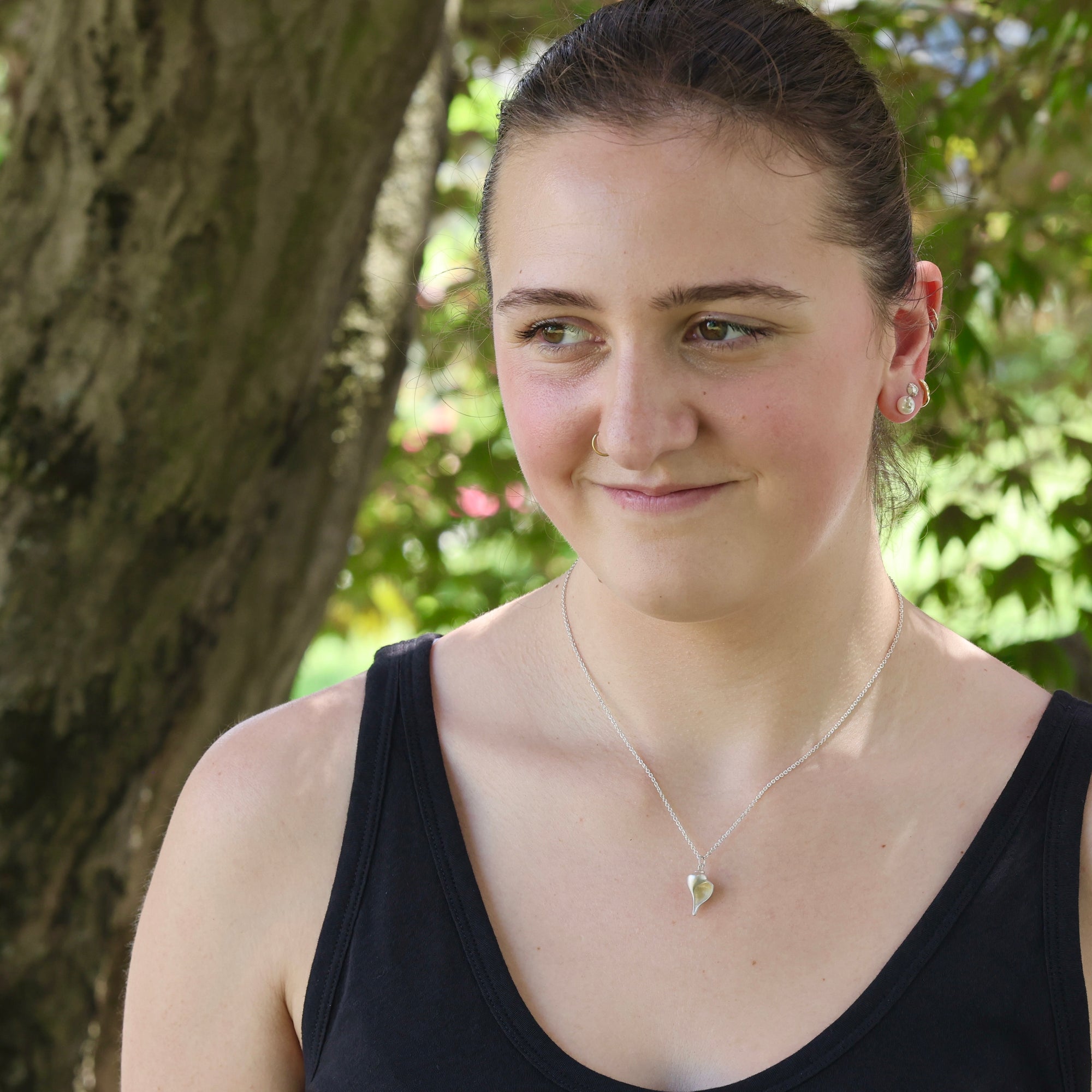 Woman wearing a black tank top with a necklace, standing outdoors with greenery in the background