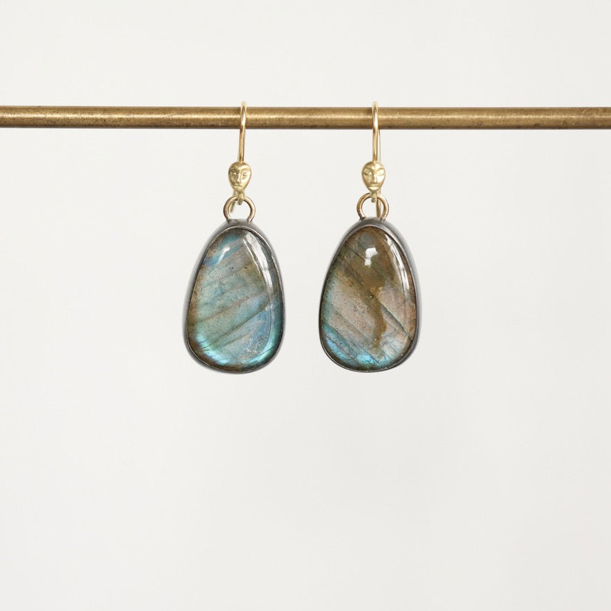 A pair of teardrop-shaped labradorite dangle earrings with gold hooks, hanging from a gold horizontal bar against a clean white background.