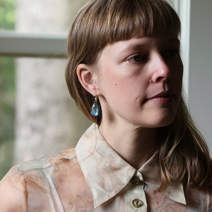 A close-up portrait of a woman with brown hair and bangs wearing a sheer beige patterned blouse and a single dangling blue teardrop earring, posed in front of a softly blurred window.