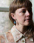 A close-up portrait of a woman with brown hair and bangs wearing a sheer beige patterned blouse and a single dangling blue teardrop earring, posed in front of a softly blurred window.