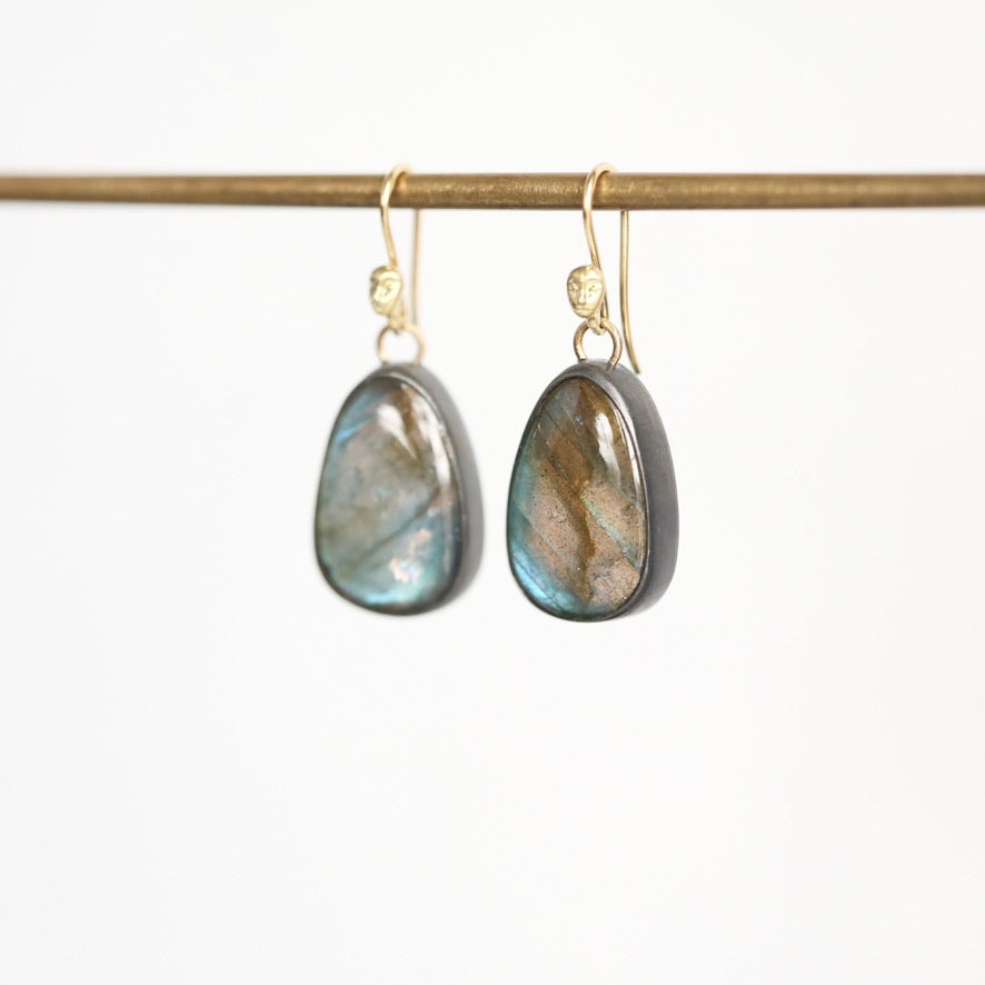 A pair of teardrop-shaped labradorite drop earrings with gold hooks, hanging from a gold rod against a clean white background.