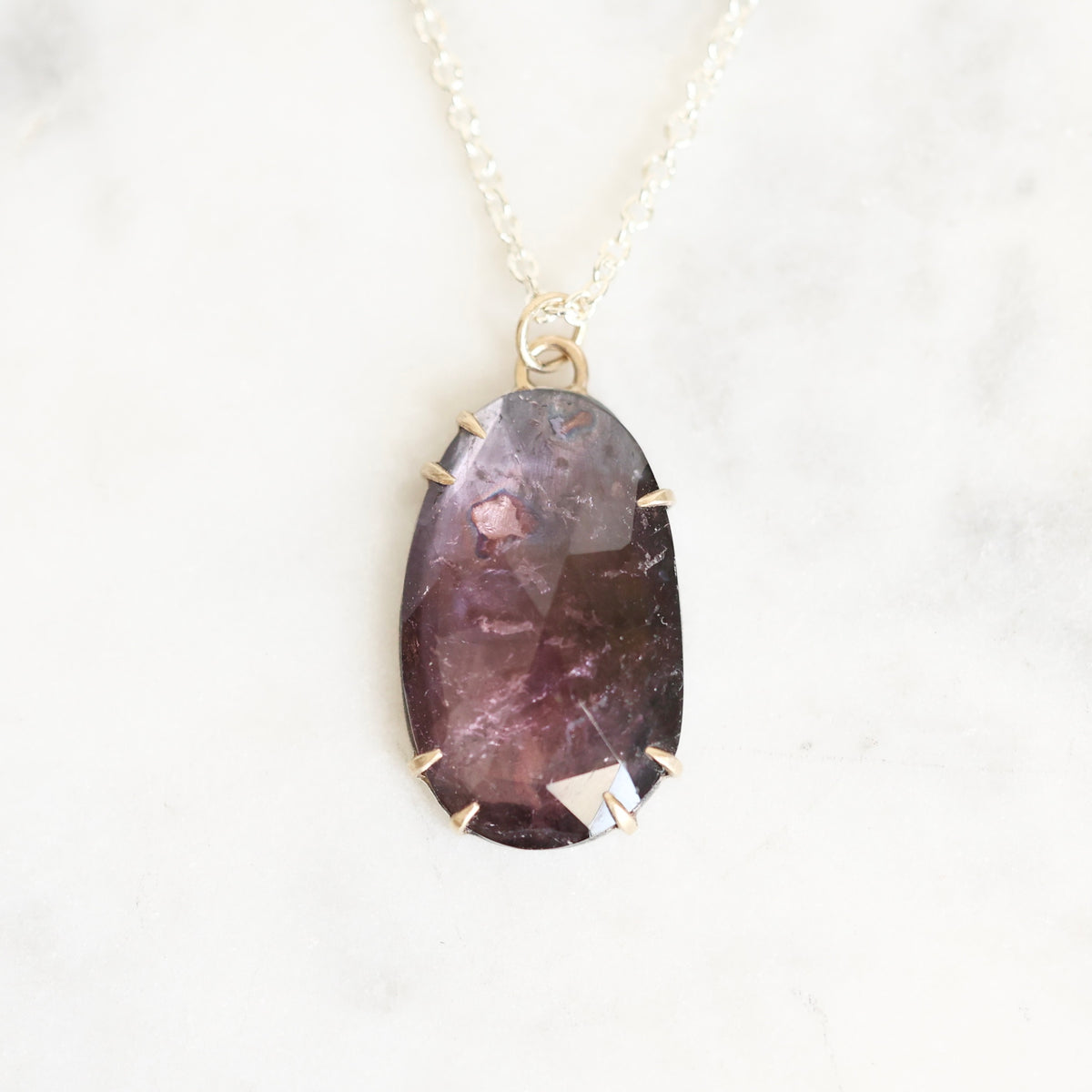 A teardrop-shaped purple gemstone pendant with gold prongs, hanging from a delicate gold chain on a white marble background.