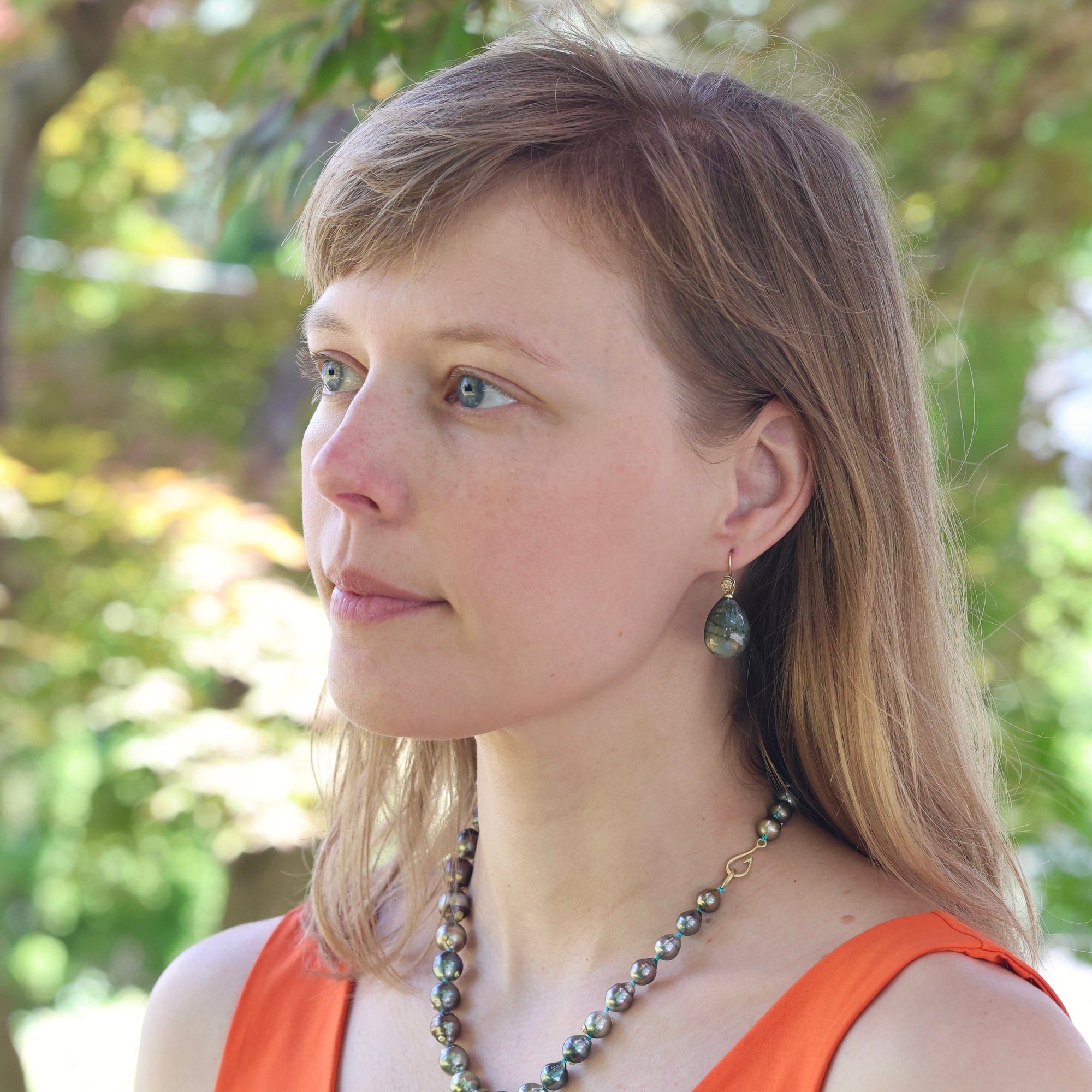 Woman with long blonde hair wearing an orange top and a necklace, standing outdoors with greenery in the background.