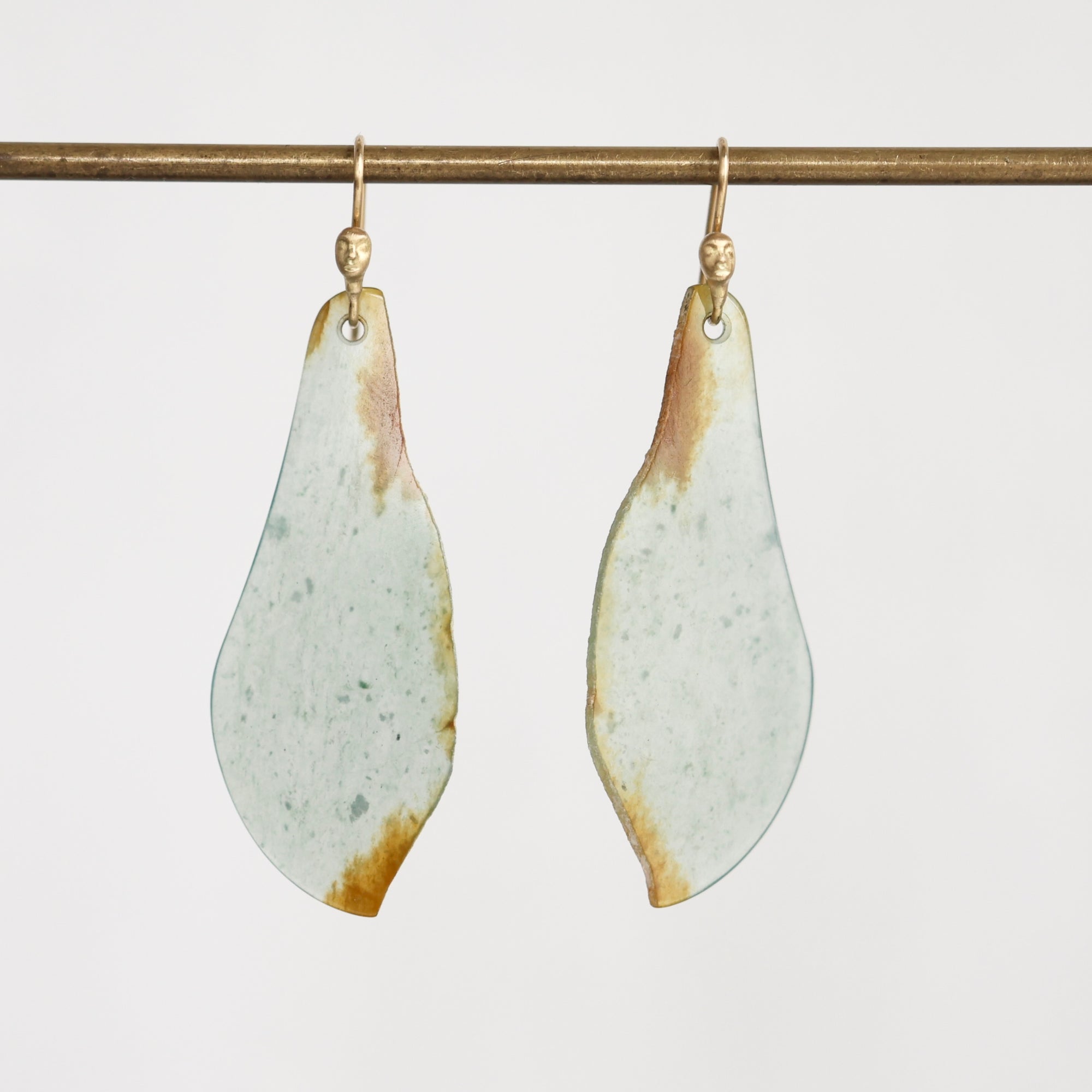 A pair of teardrop-shaped drop earrings with a light sage green and white marbled surface and natural brown edges, hanging from gold hooks on a horizontal brass rod against a plain white background.