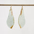 A pair of teardrop-shaped drop earrings with a light sage green and white marbled surface and natural brown edges, hanging from gold hooks on a horizontal brass rod against a plain white background.