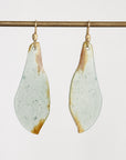 A pair of teardrop-shaped drop earrings with a light sage green and white marbled surface and natural brown edges, hanging from gold hooks on a horizontal brass rod against a plain white background.