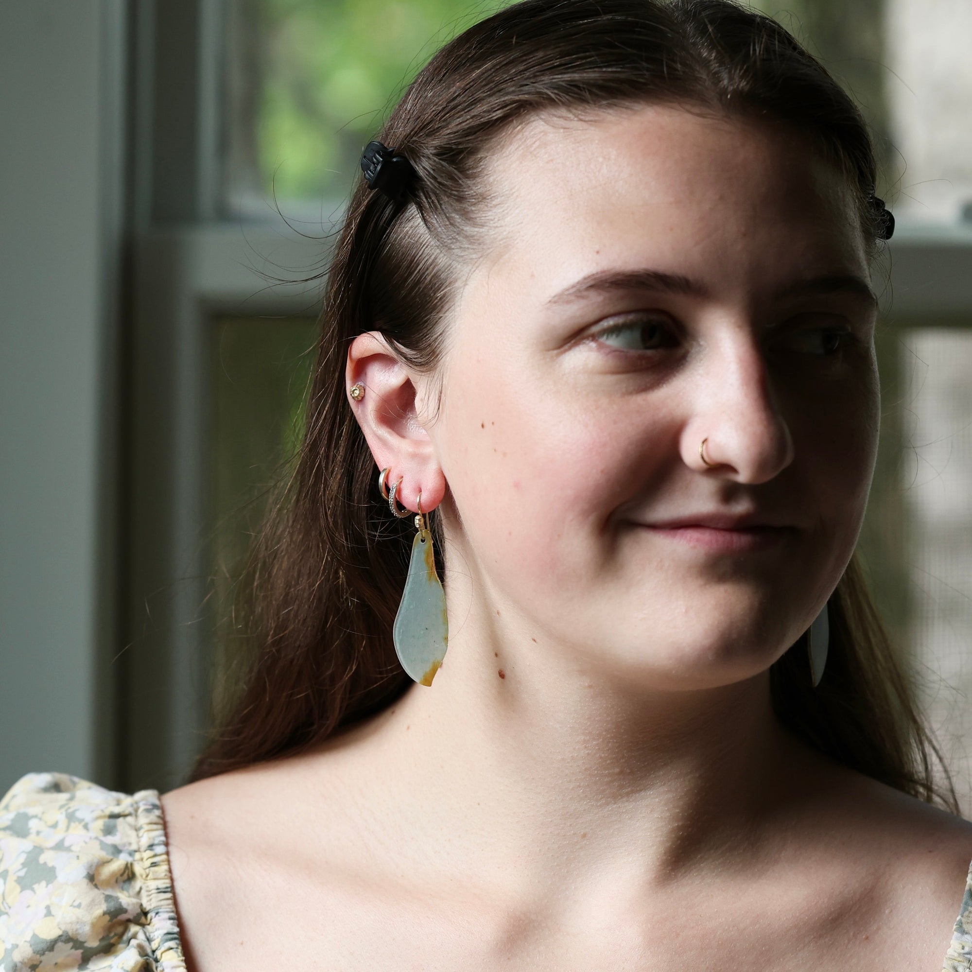 A close-up portrait of a woman wearing a pair of long, teardrop-shaped dangle earrings with a translucent light blue-green center and small gold-toned accents, set against a softly blurred window background.