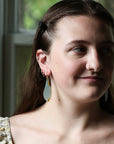 A close-up portrait of a woman wearing a pair of long, teardrop-shaped dangle earrings with a translucent light blue-green center and small gold-toned accents, set against a softly blurred window background.
