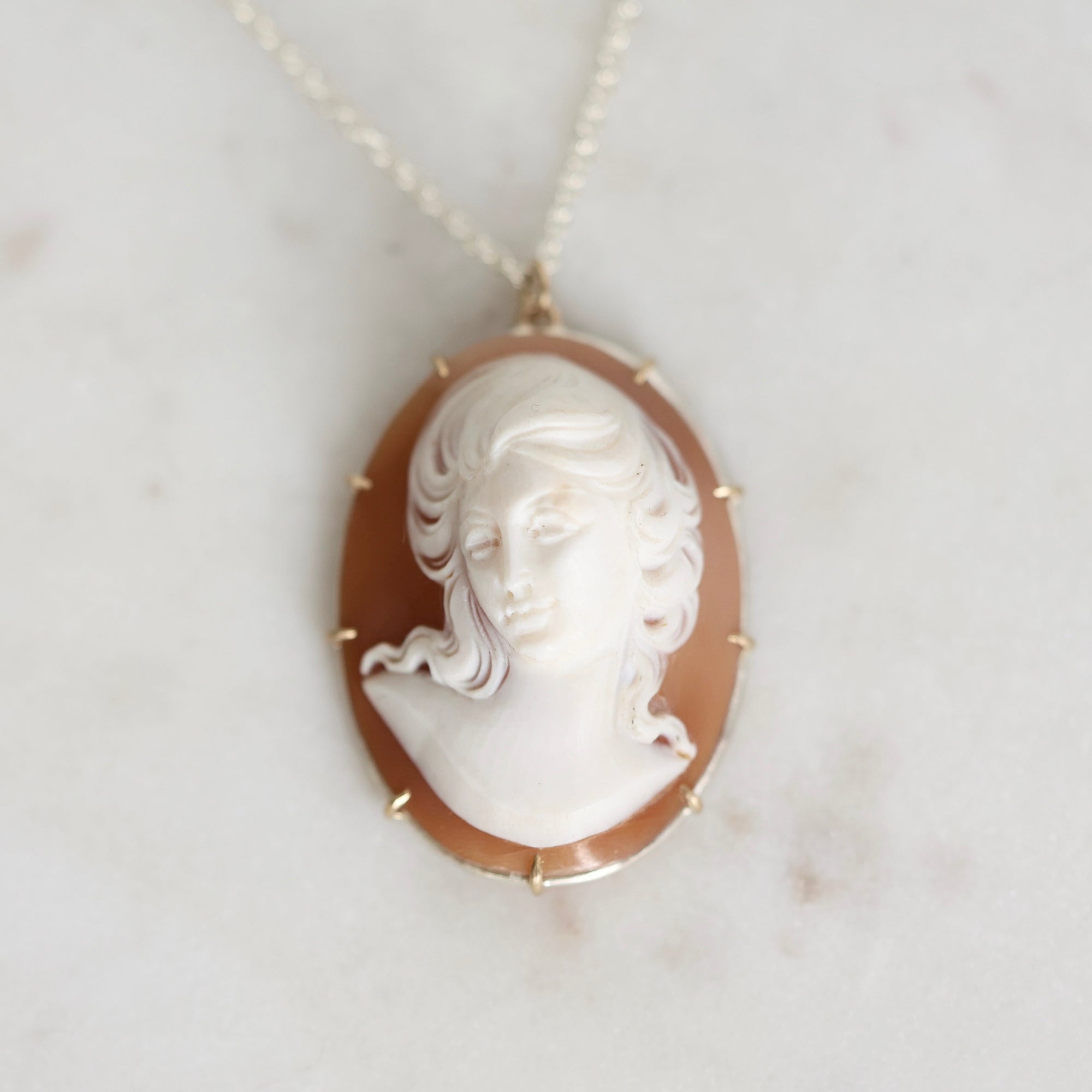 An oval pendant necklace featuring a carved white classical bust on a warm brown background, set in a gold-toned frame and hanging from a delicate chain on a light marble surface.