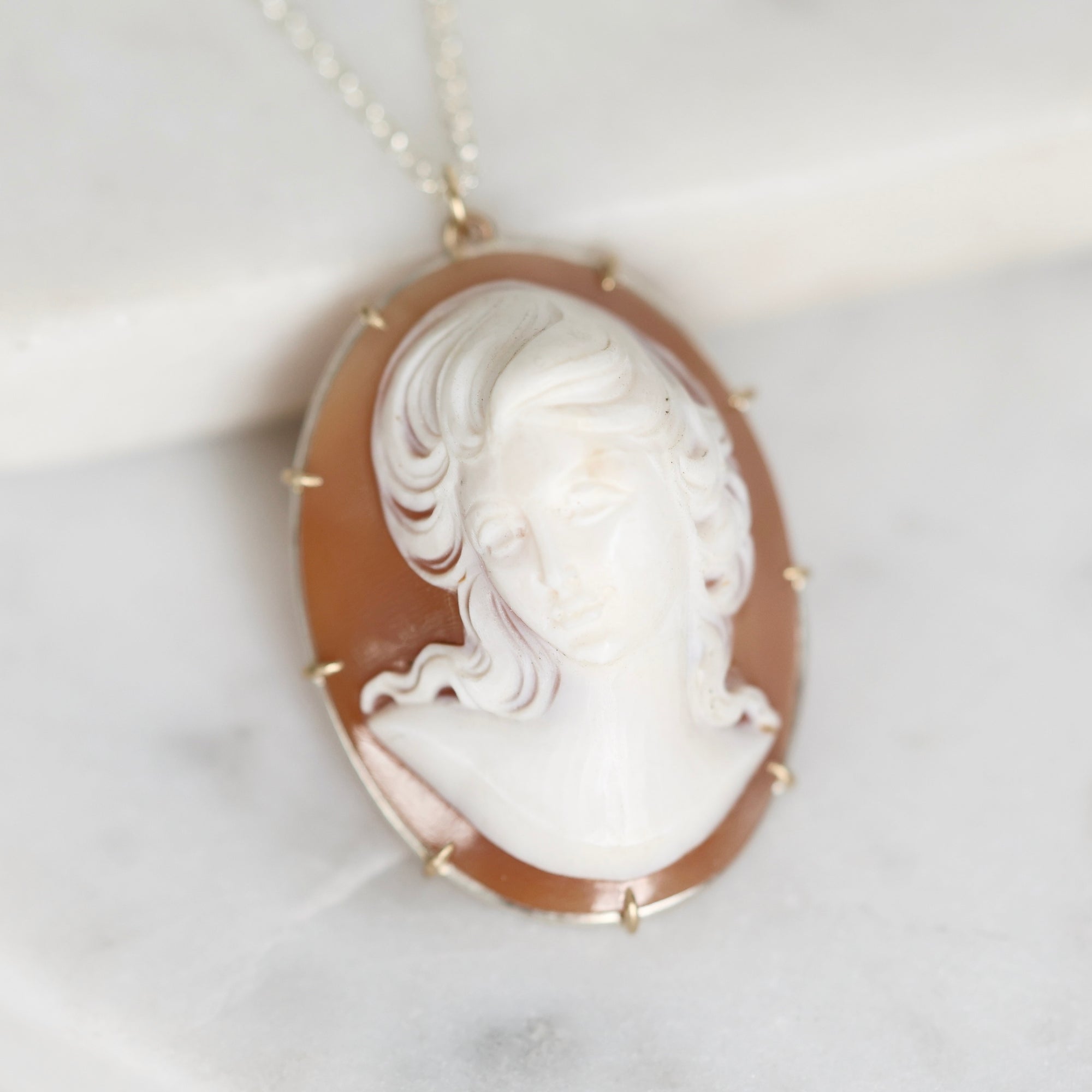 An oval pendant necklace featuring a carved white classical bust on a warm brown shell-like background, set in a gold-toned bezel with small gold spikes, hanging from a delicate chain on a light marble surface.