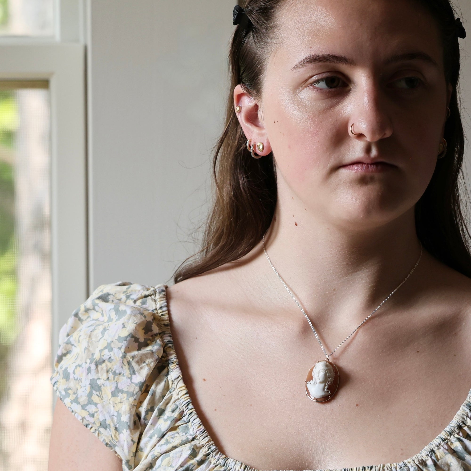 A close-up lifestyle shot of a woman wearing a silver chain necklace with a carved shell pendant, paired with a floral-patterned puff-sleeve top, set against a soft-focus indoor background.