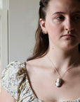 A close-up lifestyle shot of a woman wearing a silver chain necklace with a carved shell pendant, paired with a floral-patterned puff-sleeve top, set against a soft-focus indoor background.