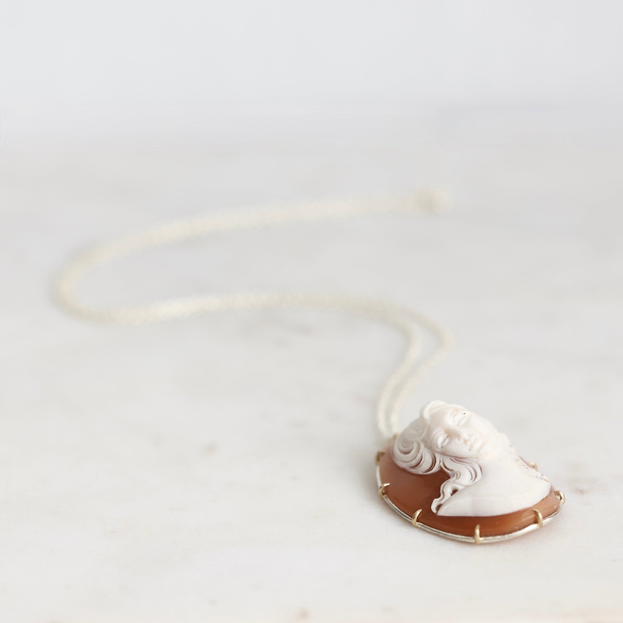 A classic Italian-style shell-shaped cameo pendant necklace featuring a white carved face on a warm brown background, set in gold-toned metal and suspended from a delicate white chain, displayed on a light marble surface.