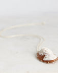 A classic Italian-style shell-shaped cameo pendant necklace featuring a white carved face on a warm brown background, set in gold-toned metal and suspended from a delicate white chain, displayed on a light marble surface.