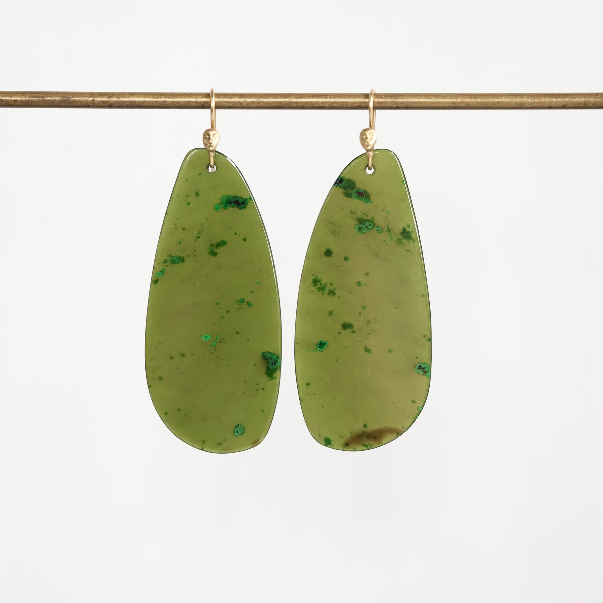 A pair of large, teardrop-shaped olive green resin drop earrings with green mineral inclusions, hanging from gold-tone fishhook ear wires on a horizontal brass rod against a white background.