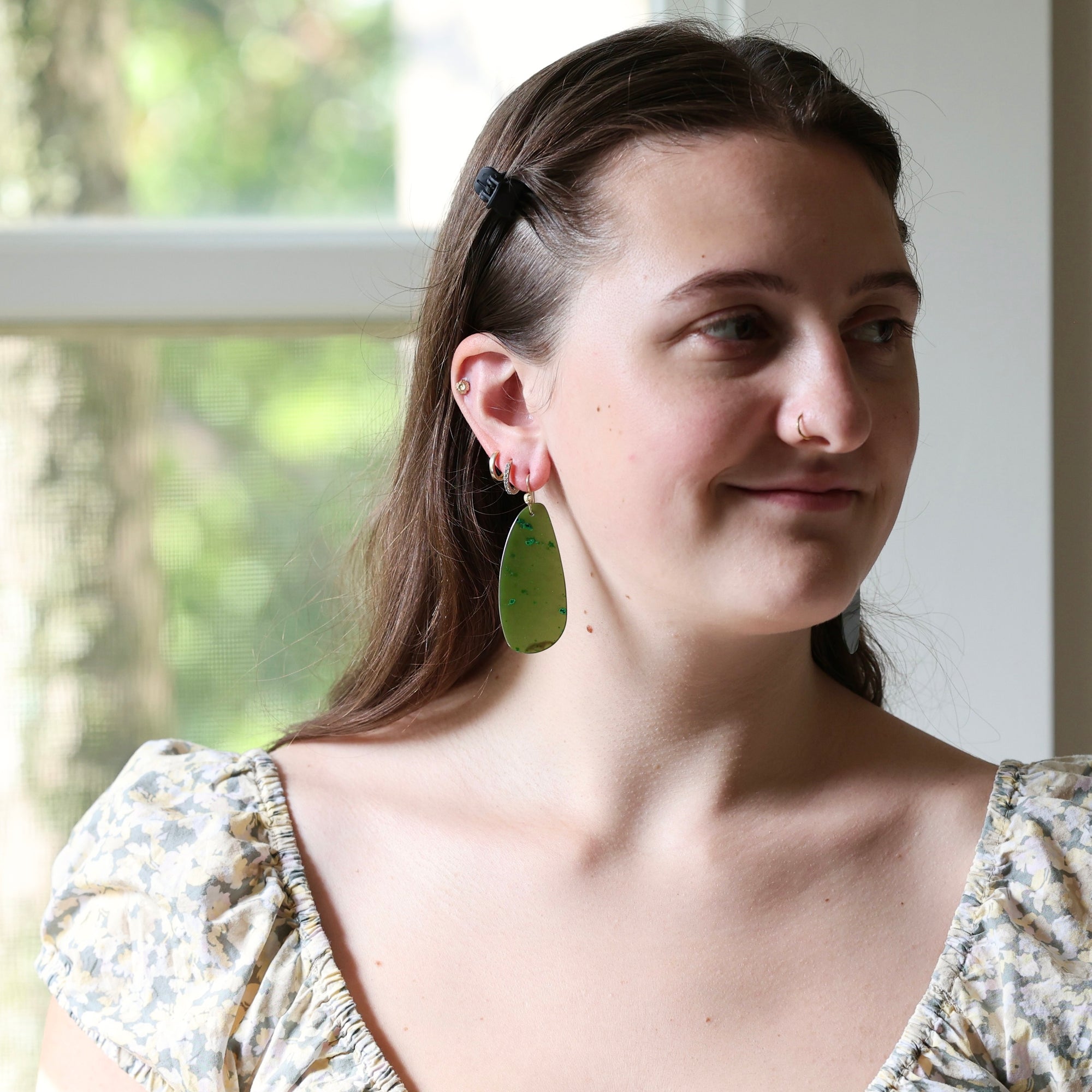 A woman wearing a large, organic-shaped olive green drop earring, shown in a close-up portrait against a softly blurred window background.
