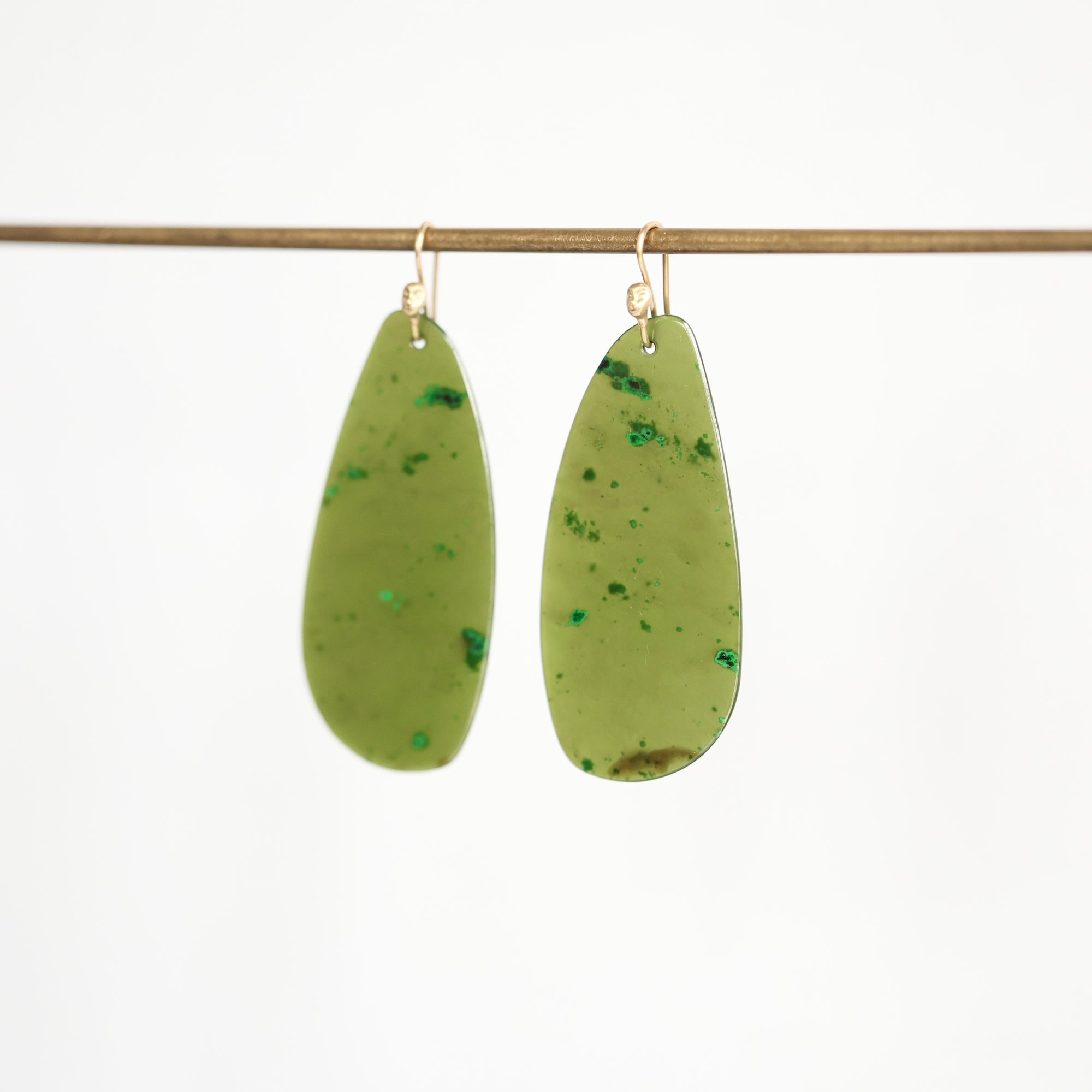 A pair of teardrop-shaped green resin earrings with tiny green inclusions and gold-tone hooks, hanging from a thin horizontal rod against a plain white background.